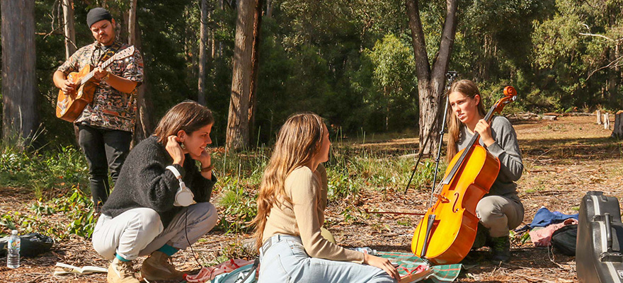 Students sitting on rugs in the forest listening to musicians play instruments