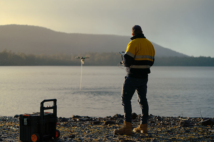 Person in high-vis standing on lake's shore, controlling a drone that can be seen in the distance collecting a water sample from the lake