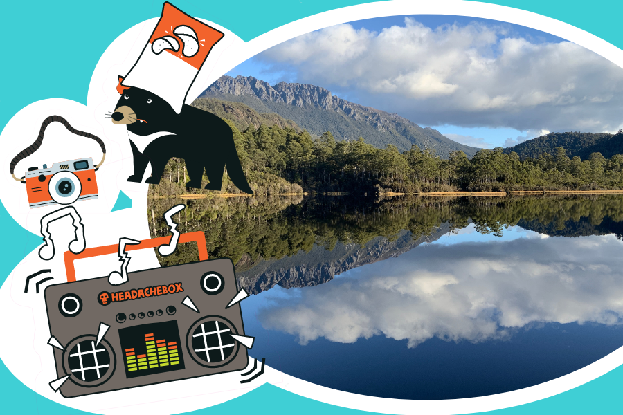 Lake Rosebery on Tasmania's West Coast, with mountains and trees behind it. The clouds and blue skies are reflecting in the lake