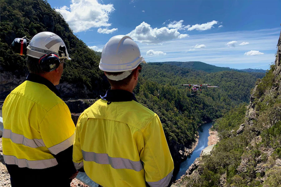 Two people wearing high-vis and hard hats controlling a drone flying out in front of them, standing on Cethana Dam