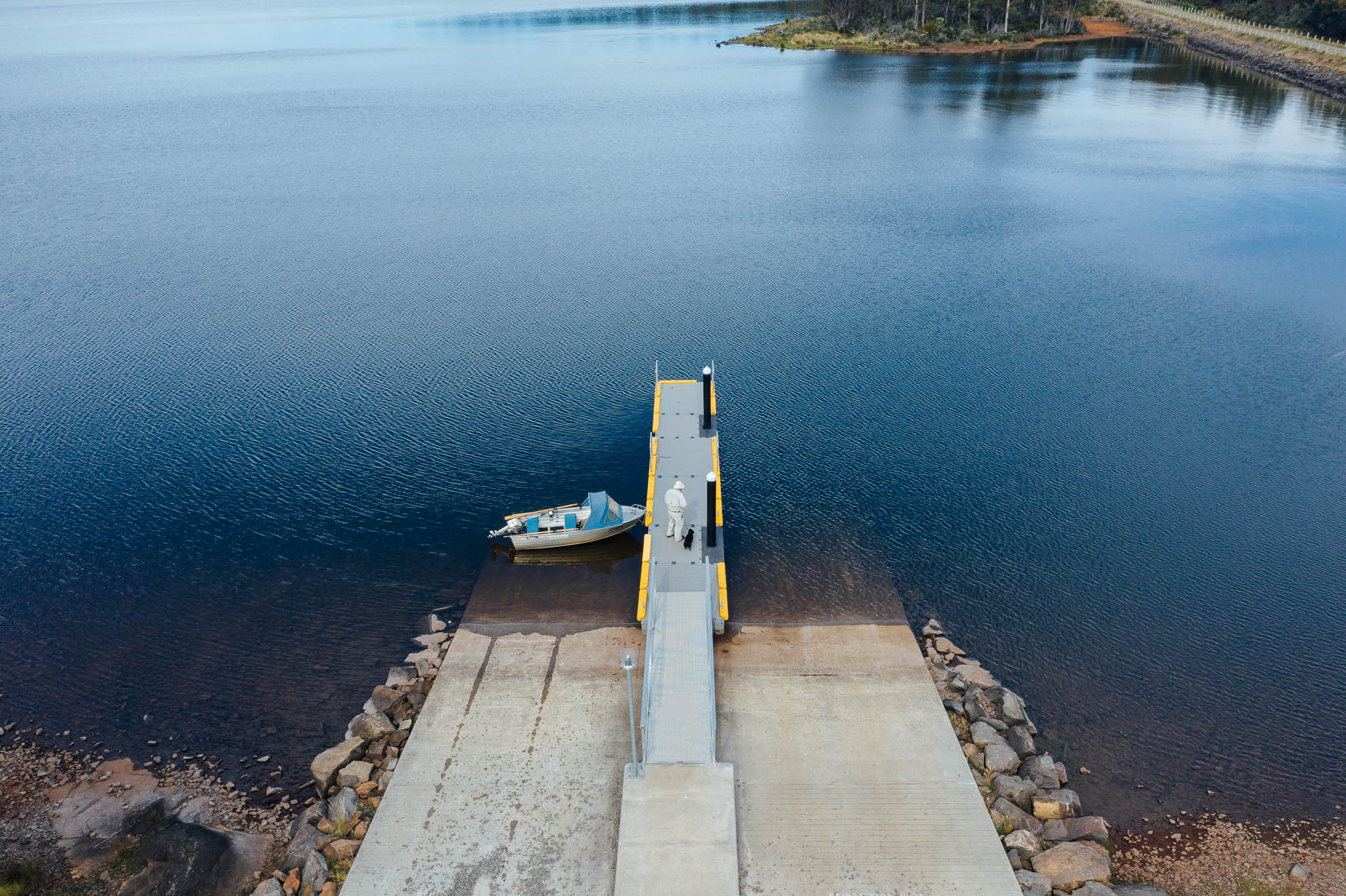 Looking down on Bronte Lagoon boat ramp, extending out from the shoreline into the clear blue lagoon. A man stands on the boat ramp, with his boat tied to the left-hand side.