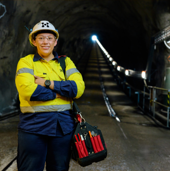 Leah is wearing a high-vis shirt, and sitting on a log facing the camera. She is holding a safety helmet. In the background is grass and trees.
