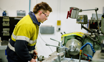 Xavier is wearing a high-vis shirt, and smiling for the camera. On his left is a large turbine part, and behind him is a workshop interior.