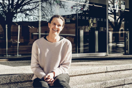 Carolyn Maxwell, sitting on the front steps of the Hydro Tasmania head office, smiling for a photograph. Behind her are windows reflecting back towards trees and blue skies.