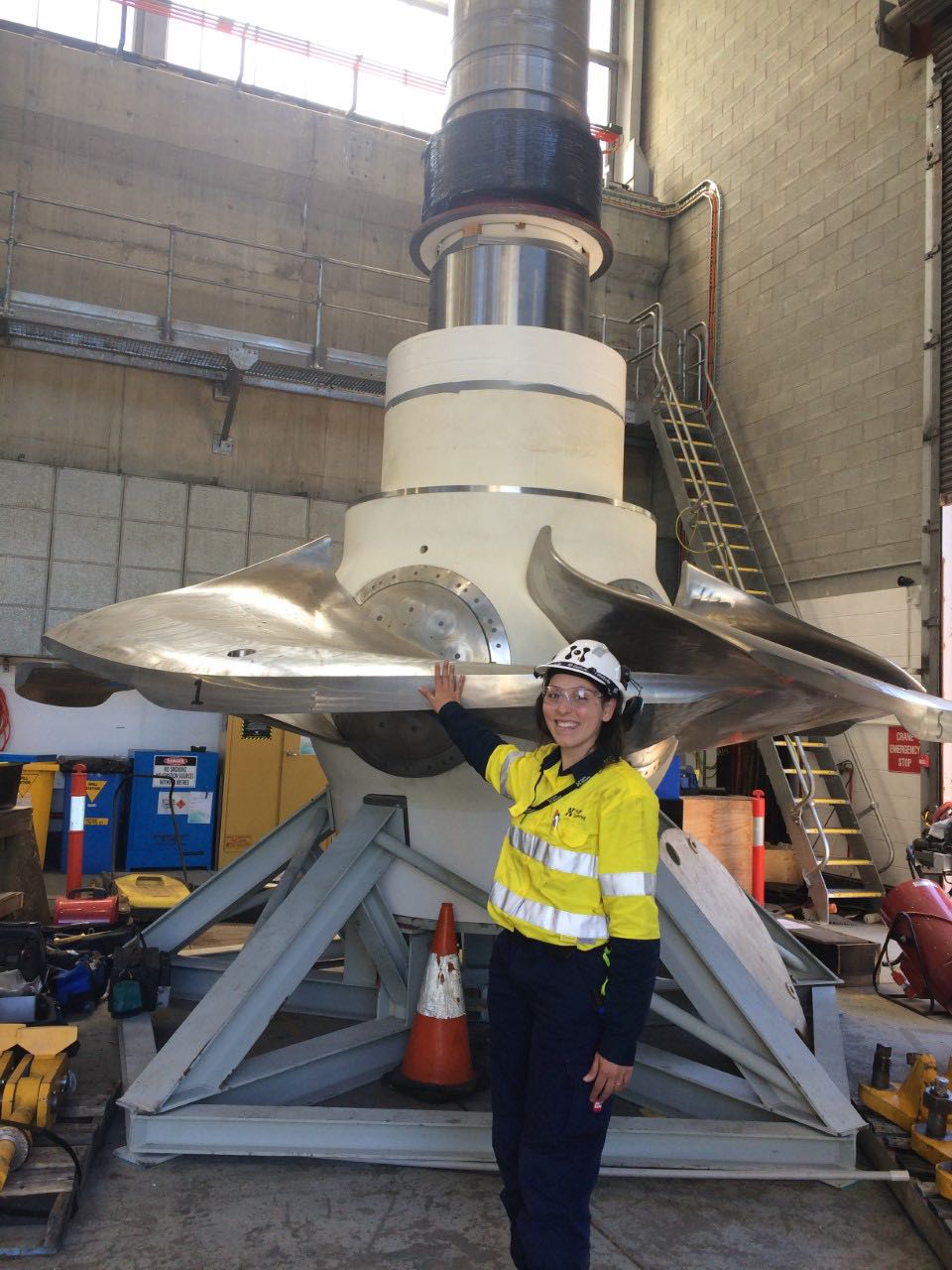 A woman wearing high vis and personal protective equipment standing in front of a turbine three times her height