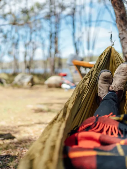 Person lying in a hammock outdoors, seen from their perspective with boots and a blanket visible, surrounded by trees, a camp chair, and a boat in the background.