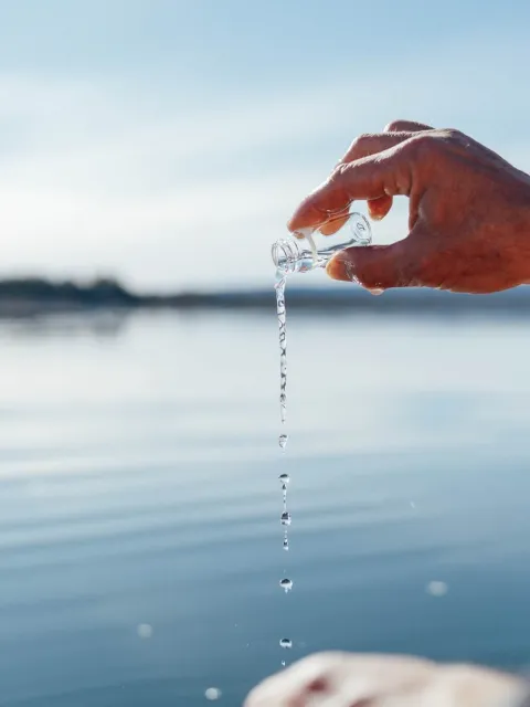 Hand holding a small glass vial, pouring water into a calm lake under a clear sky