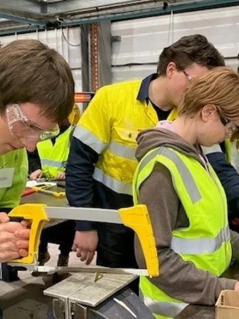 Group of students in high-visibility safety vests working with tools in an industrial workshop.
