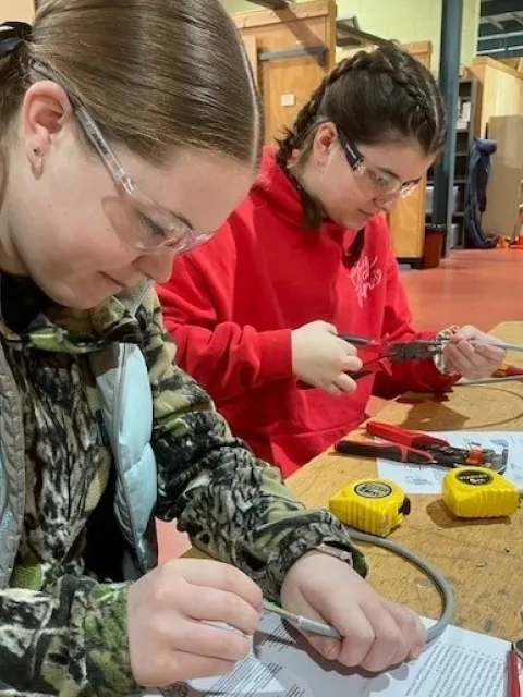 Two students working at a table with wires, pliers, and measuring tapes in a workshop setting.