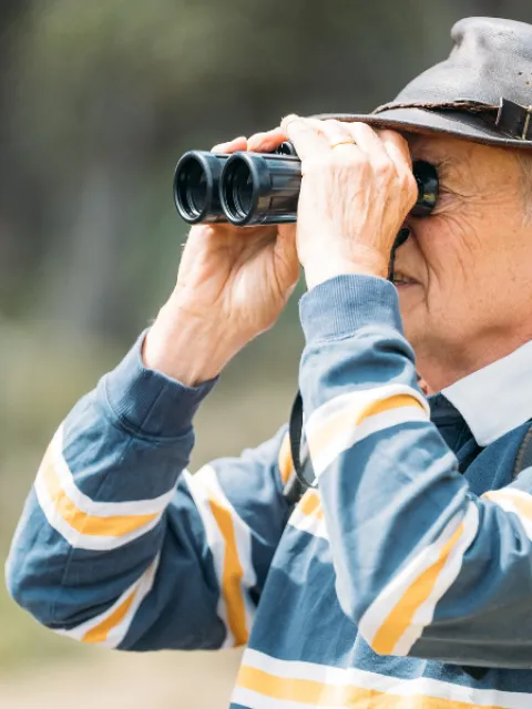 Person outdoors using binoculars, wearing a striped sweater and a wide-brimmed hat