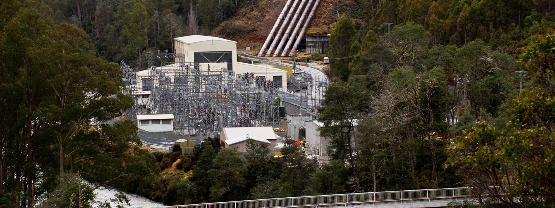 Hydropower station at the base of a forested hillside with large silver pipes descending toward a river and a bridge crossing in the foreground.